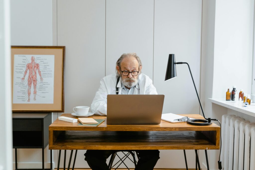 Elderly doctor in office using a laptop with medical books and anatomical chart.